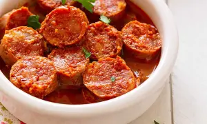 Chorizos a la sidra jugosos y dorados en una salsa rojiza, servidos en un cuenco blanco.