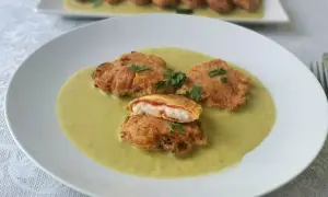 Pimientos piquillo rellenos rebozados, servidos en una cremosa salsa verde. Uno está cortado, mostrando su interior.