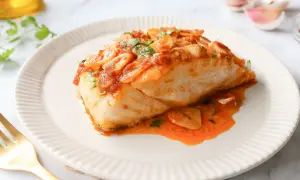 Bacalao a la llauna jugoso, cubierto de salsa de tomate y ajo laminado, servido en plato blanco con tenedor dorado.