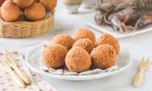 Croquetas de gambas doradas y crujientes en un plato blanco, con más en una cesta de mimbre al fondo y gambas frescas.