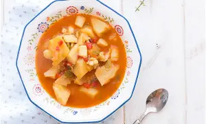Un plato de sepia con patatas, pimientos y trozos de tomate en una salsa rojiza, servido en un cuenco decorado.