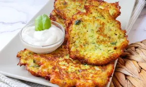 Tortitas de calabacín doradas y crujientes, servidas con una cremosa salsa y decoradas con una hoja de albahaca.