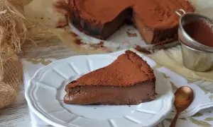 Una rebanada de tarta de chocolate, cubierta de cacao en polvo, reposa en un plato blanco. El postre parece cremoso y delicioso.