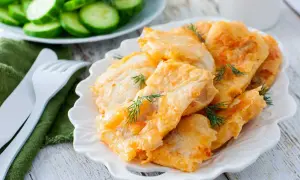 Delicioso cachopo de merluza rebozado, servido en plato blanco con eneldo fresco. Acompañado de rodajas de pepino.