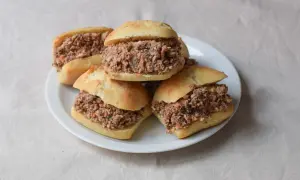 Cinco montaditos de pringa, rellenos de carne desmenuzada y especias, apilados en un plato blanco sobre un mantel de lino.