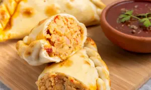 Empanada de pollo recién horneada, cortada por la mitad, revela un relleno jugoso. A su lado, un cuenco de salsa roja con perejil.