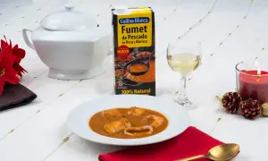 Caldereta de pescado servida en plato blanco, acompañada de fumet de pescado y marisco, vino blanco y vela encendida.