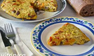 Deliciosa tortilla de berenjena con pimientos y calabaza, servida en un plato decorado. Acompañada de pan rústico y cubiertos.