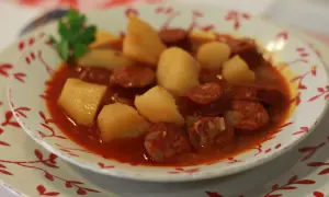 Un plato humeante de patatas a la riojana, con trozos de chorizo y patata en una salsa rojiza, decorado con una ramita de perejil.
