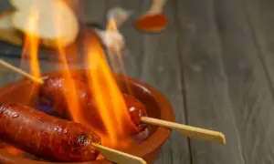 Chorizo al infierno, dos salchichas en brochetas ardiendo en una cazuela de barro, con pan y pimentón al fondo.