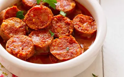 Chorizos a la sidra jugosos y dorados en una salsa rojiza, servidos en un cuenco blanco.