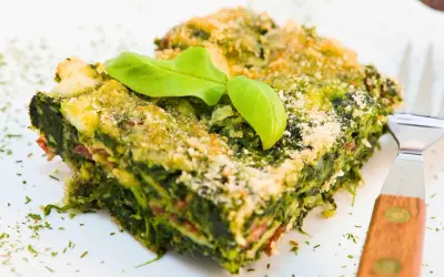 Porción de lasaña verde con capas de pencas de acelgas, queso y una hoja de albahaca encima.