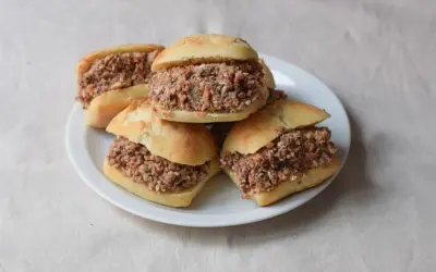Cinco montaditos de pringa, rellenos de carne desmenuzada y especias, apilados en un plato blanco sobre un mantel de lino.
