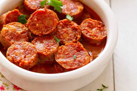 Chorizos a la sidra jugosos y dorados en una salsa rojiza, servidos en un cuenco blanco.