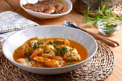 Deliciosa cazuela de pescado con gambas y trozos de pescado blanco en una salsa rojiza, adornada con perejil fresco.