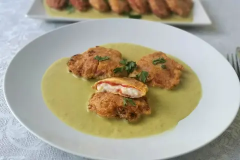 Pimientos piquillo rellenos rebozados, servidos en una cremosa salsa verde. Uno está cortado, mostrando su interior.