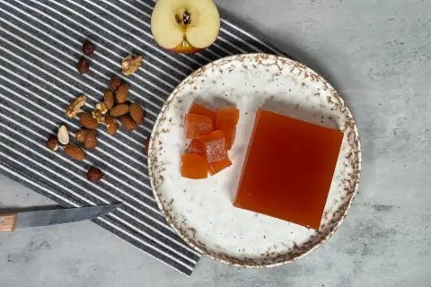 Un trozo de dulce de manzana y cubos cortados en un plato, junto a frutos secos y una manzana a medio cortar.