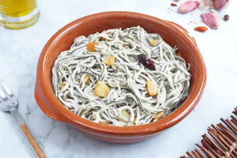 Un cuenco de barro con gulas al ajillo, salteadas con ajo y guindilla. Se ve un tenedor y una botella de aceite de oliva.