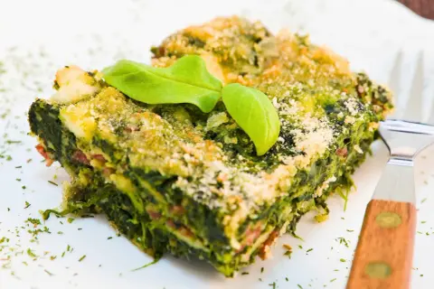 Porción de lasaña verde con capas de pencas de acelgas, queso y una hoja de albahaca encima.