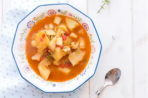 Un plato de sepia con patatas, pimientos y trozos de tomate en una salsa rojiza, servido en un cuenco decorado.