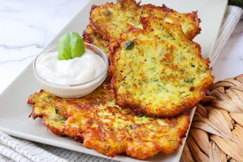 Tortitas de calabacín doradas y crujientes, servidas con una cremosa salsa y decoradas con una hoja de albahaca.