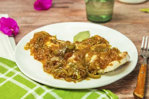 Un plato de urta a la roteña, con pescado blanco cubierto de cebolla y pimientos pochados, servido en una mesa de madera.