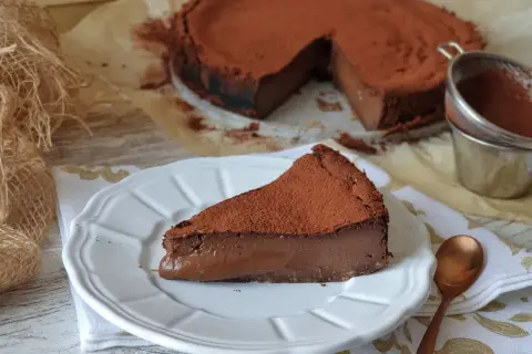 Una rebanada de tarta de chocolate, cubierta de cacao en polvo, reposa en un plato blanco. El postre parece cremoso y delicioso.