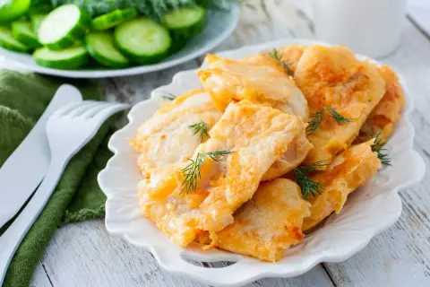Delicioso cachopo de merluza rebozado, servido en plato blanco con eneldo fresco. Acompañado de rodajas de pepino.