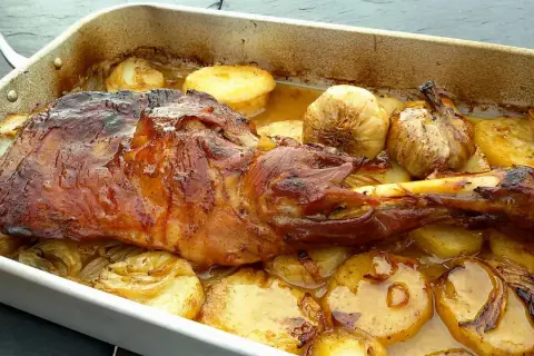 Paletilla de cordero lechal al horno estilo segoviano, dorada y jugosa, acompañada de patatas y ajos asados en su jugo.