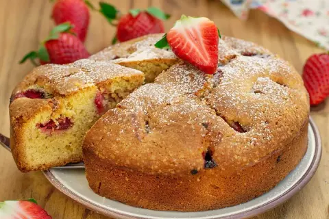 Delicioso bizcocho de fresa, espolvoreado con azúcar glas y decorado con una fresa fresca. Se ve una porción cortada, revelando trozos de fresa en su interior.