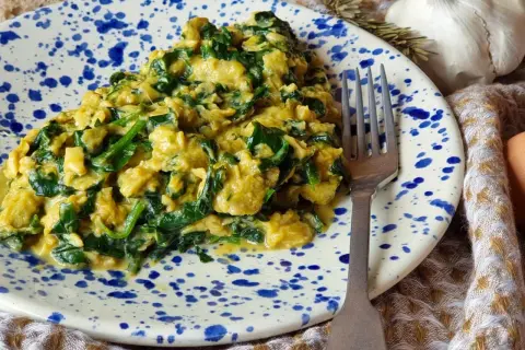 Un delicioso revuelto de espinacas cremoso, servido en un plato con salpicaduras azules. Un tenedor plateado descansa a un lado, listo para disfrutar.