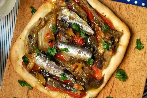 Deliciosa coca de verduras con sardinas y perejil fresco, servida sobre tabla de madera.