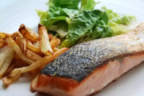 Un delicioso salmón a la plancha con piel crujiente, acompañado de patatas fritas doradas y una fresca ensalada verde.