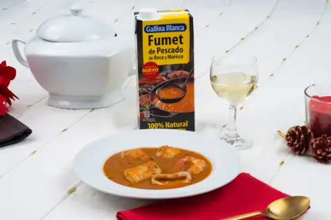 Caldereta de pescado servida en plato blanco, acompañada de fumet de pescado y marisco, vino blanco y vela encendida.