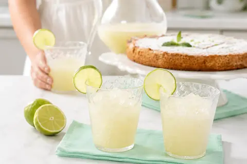 Refrescante granizado de limón servido en vasos con rodajas de lima, junto a limones frescos y un pastel.