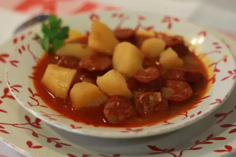 Un plato humeante de patatas a la riojana, con trozos de chorizo y patata en una salsa rojiza, decorado con una ramita de perejil.