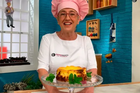 Eva Arguiñano sonríe con su delantal blanco y un gorro rosa, presentando una deliciosa tarta de queso.