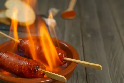 Chorizo al infierno, dos salchichas en brochetas ardiendo en una cazuela de barro, con pan y pimentón al fondo.