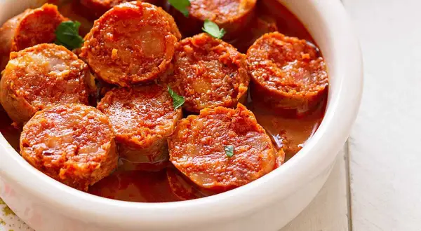 Chorizos a la sidra jugosos y dorados en una salsa rojiza, servidos en un cuenco blanco.