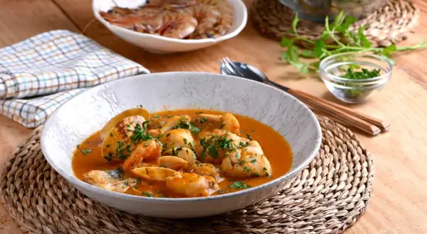 Deliciosa cazuela de pescado con gambas y trozos de pescado blanco en una salsa rojiza, adornada con perejil fresco.