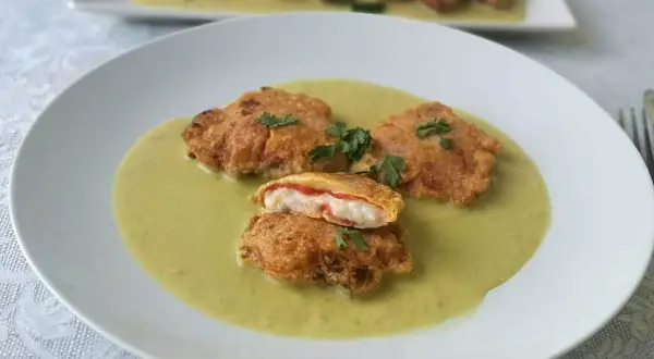 Pimientos piquillo rellenos rebozados, servidos en una cremosa salsa verde. Uno está cortado, mostrando su interior.