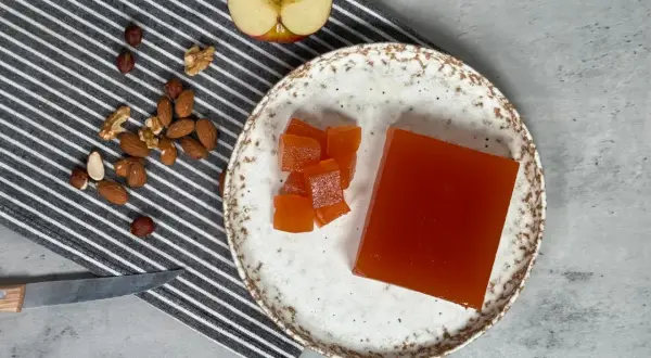 Un trozo de dulce de manzana y cubos cortados en un plato, junto a frutos secos y una manzana a medio cortar.