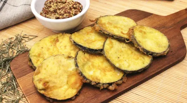 Berenjenas rebozadas con harina y huevo, doradas y crujientes, servidas en tabla de madera con un cuenco de mostaza.