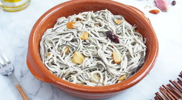 Un cuenco de barro con gulas al ajillo, salteadas con ajo y guindilla. Se ve un tenedor y una botella de aceite de oliva.