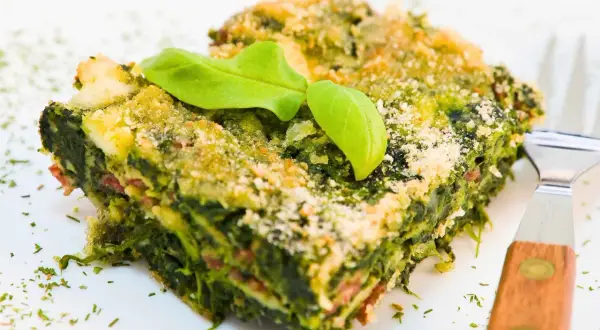 Porción de lasaña verde con capas de pencas de acelgas, queso y una hoja de albahaca encima.