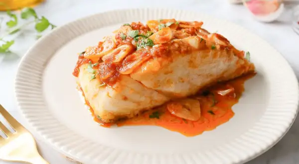 Bacalao a la llauna jugoso, cubierto de salsa de tomate y ajo laminado, servido en plato blanco con tenedor dorado.