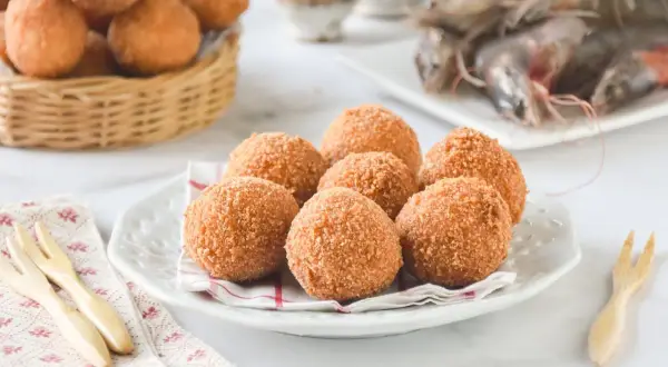 Croquetas de gambas doradas y crujientes en un plato blanco, con más en una cesta de mimbre al fondo y gambas frescas.