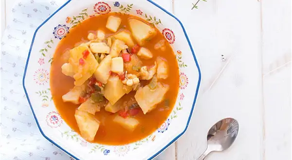Un plato de sepia con patatas, pimientos y trozos de tomate en una salsa rojiza, servido en un cuenco decorado.