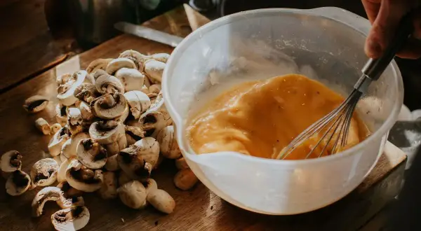 Un bol con huevos batidos y champiñones cortados en una tabla de madera, listos para un delicioso revuelto de setas.