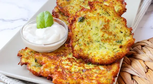 Tortitas de calabacín doradas y crujientes, servidas con una cremosa salsa y decoradas con una hoja de albahaca.